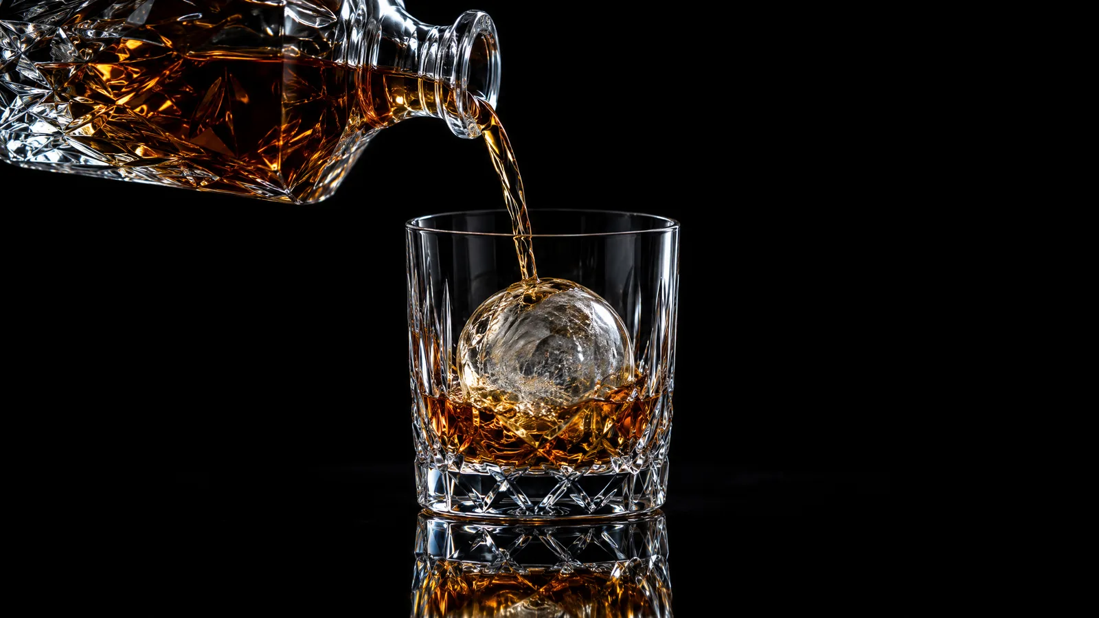 Amber brandy pouring from a crystal decanter over a sphere of ice in a heavy tumbler