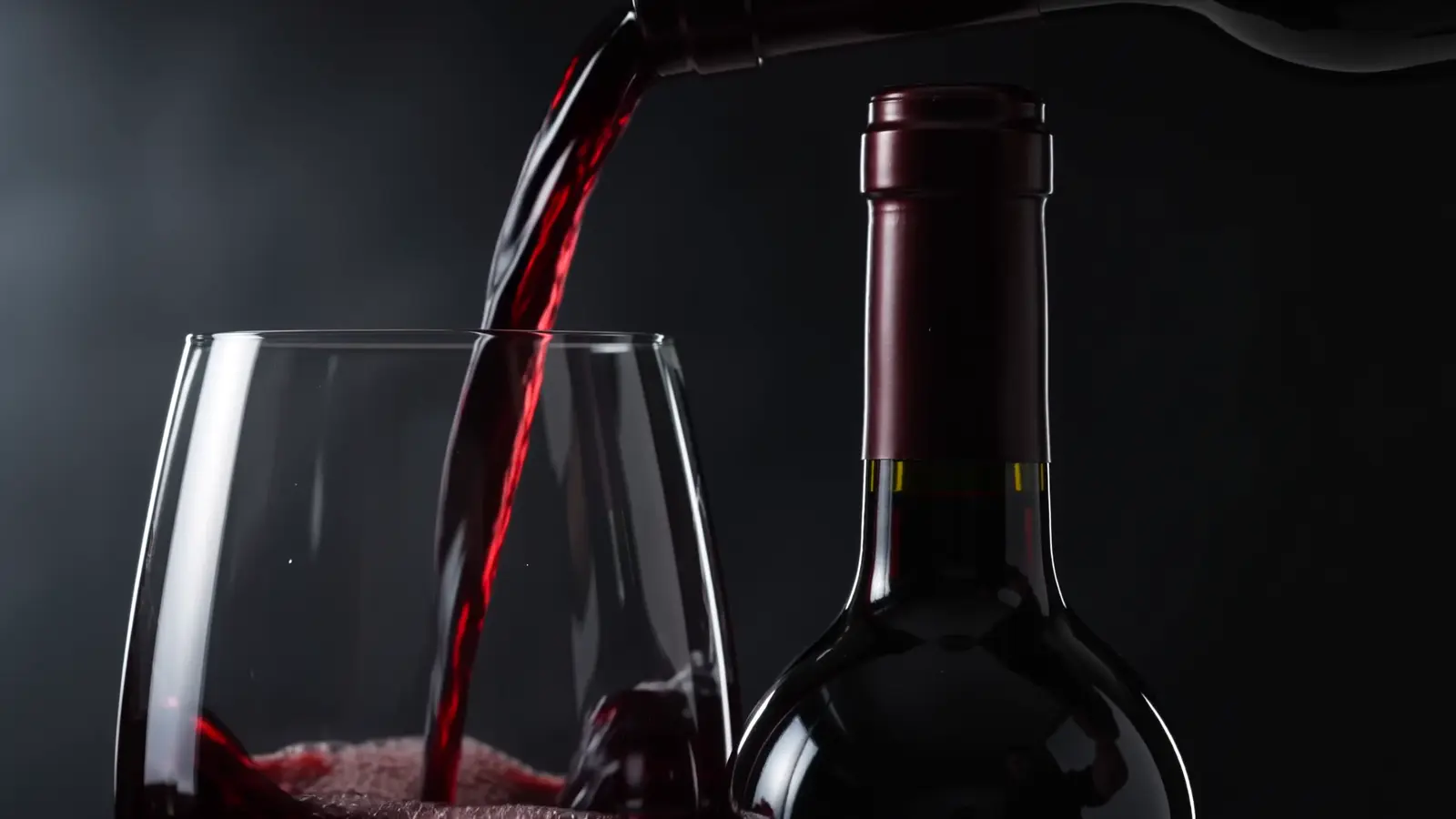 Red wine being poured into a crystal glass beside a sealed, foil-capped bottle in low silver light