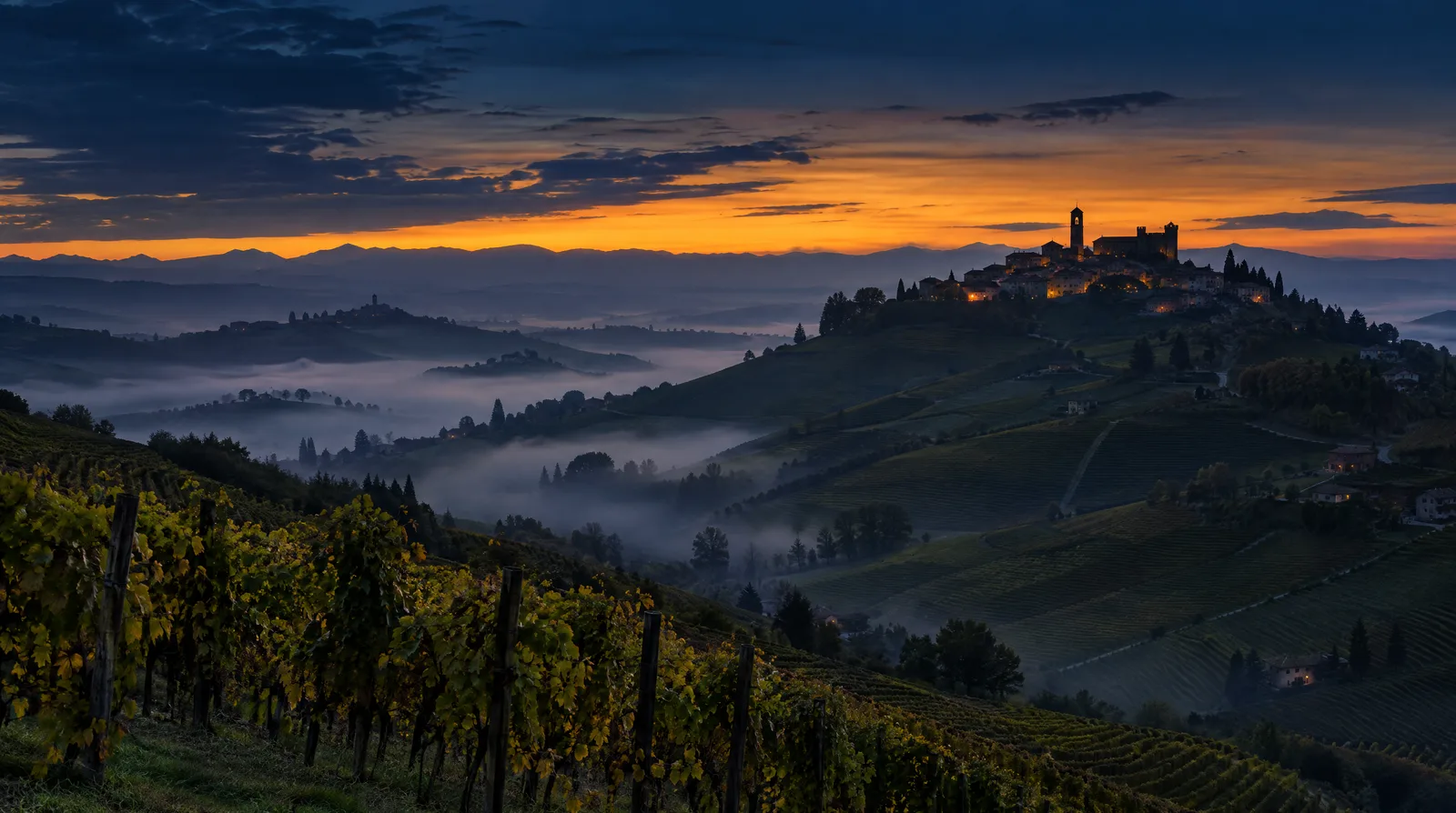The vineyard hills of Piedmont's Langhe at dusk with a hilltop village against a deep blue sky