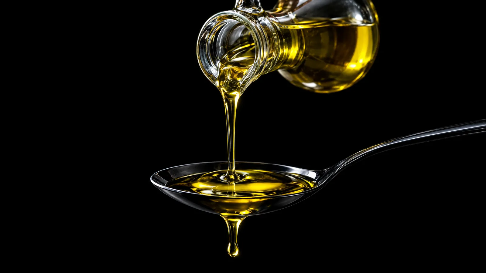A thin golden stream of olive oil pouring from a glass cruet into a polished black spoon