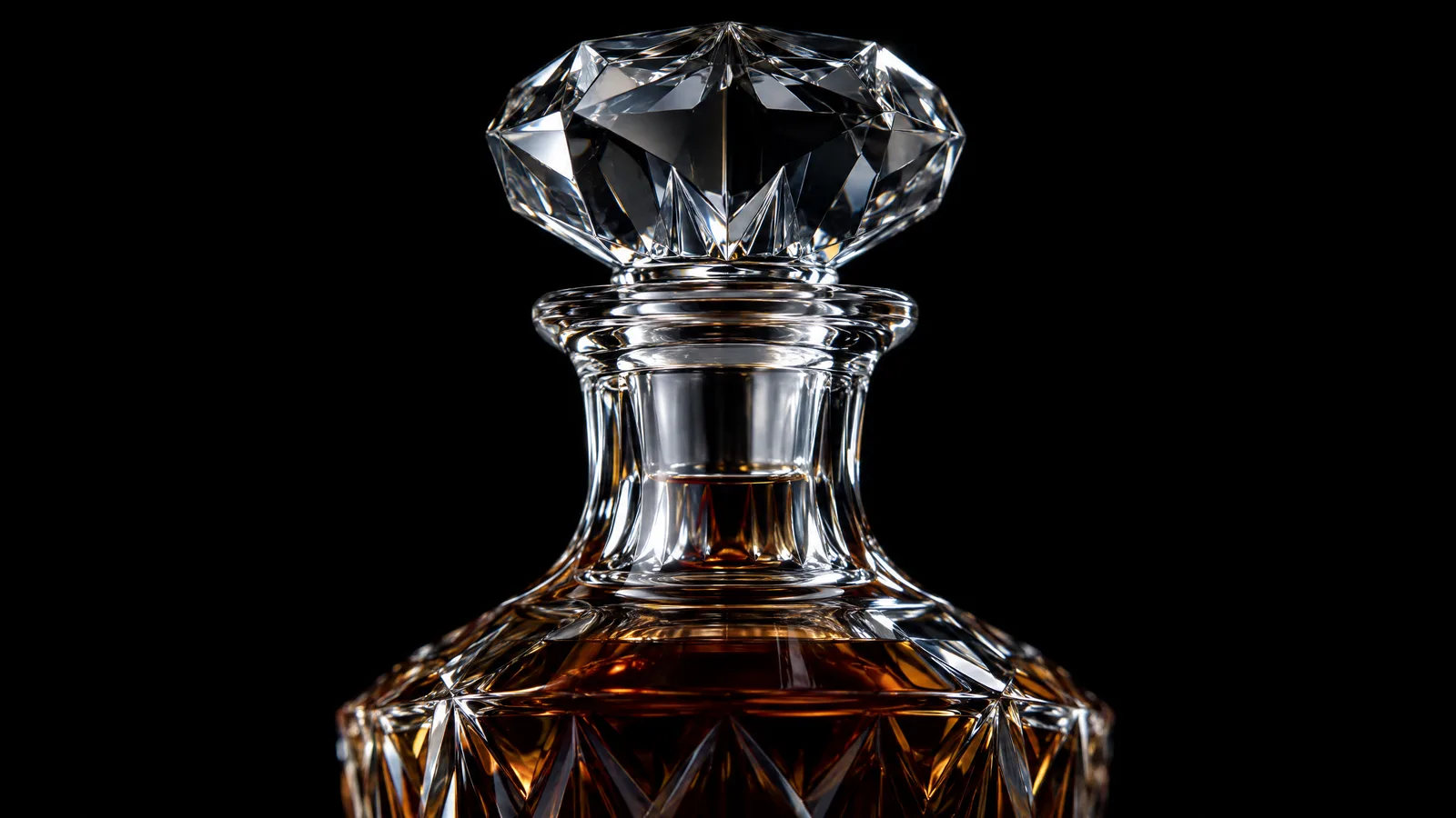 The faceted crystal stopper seated in the neck of a decanter of amber brandy, lit in silver against black
